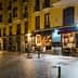 Street view of a yellow building featuring a café with outdoor seating and illuminated signage at night. - Olive Oil Times