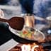 Chef using a wooden spatula to stir colorful vegetables in a frying pan over a flame. - Olive Oil Times