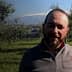 Man standing in an olive grove with olive trees in the background and mountains visible in the distance. - Olive Oil Times