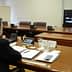 Man attending a virtual meeting with multiple participants displayed on a screen in a conference room. - Olive Oil Times