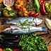 Two whole fish placed on a wooden board surrounded by various vegetables and herbs. - Olive Oil Times