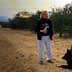 A woman holding a glass and standing next to two dogs in an olive grove. - Olive Oil Times