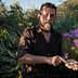 Man with a beard holding soil in his hand while standing among colorful flowers. - Olive Oil Times