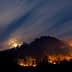 A mountain landscape with visible flames and smoke under a night sky with clouds and a full moon. - Olive Oil Times