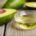 Sliced avocado with a pit next to a small glass bowl of avocado oil on a wooden surface. - Olive Oil Times