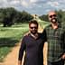 Two men posing together in an olive grove with trees and grass in the background. - Olive Oil Times