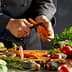 Chef peeling a carrot with various vegetables and herbs on a kitchen counter. - Olive Oil Times