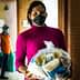 A woman wearing a pink sweater and black mask holds a bag of food items in a corridor. - Olive Oil Times