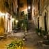 Narrow street featuring outdoor tables and potted plants, illuminated by street lamps at night. - Olive Oil Times