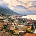 Lakeside village with houses and mountains in the background under a cloudy sky. - Olive Oil Times