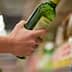 A person holding a green bottle of olive oil while reaching for another on a shelf. - Olive Oil Times