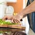 A person using a knife to chop green vegetables on a wooden cutting board in a kitchen. - Olive Oil Times