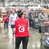 A woman in a red vest stands in a Costco store checkout area with shopping carts and customers in the background. - Olive Oil Times