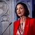 Woman in a red blazer speaking at a podium with a decorative background. - Olive Oil Times