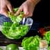 Person tearing fresh green lettuce leaves into a glass bowl for salad preparation. - Olive Oil Times