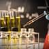A laboratory scene showing a hand in a blue glove using a pipette to transfer liquid into small glass bottles. - Olive Oil Times