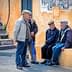Four elderly men wearing caps engaged in conversation while seated and standing near a colorful wall. - Olive Oil Times