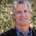 Man with gray hair and a blue shirt smiling in front of olive trees. - Olive Oil Times