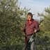 Man wearing a checkered shirt holding an olive harvesting tool in an olive grove. - Olive Oil Times