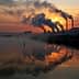 Silhouetted industrial power plant with smoke stacks against a sunset sky and water reflection. - Olive Oil Times