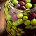 Glass jar filled with a mix of green and black olives on a wooden surface. - Olive Oil Times