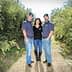 Three individuals standing together in an olive grove, wearing casual clothing and hats. - Olive Oil Times