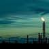 Flare stack emitting flame against a dusk sky with clouds and silhouettes of industrial structures. - Olive Oil Times