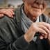 Elderly man with gray hair holding a cane, wearing a gray cardigan, with a hand on his shoulder. - Olive Oil Times