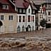 Residential buildings partially submerged in floodwaters with strong currents visible. - Olive Oil Times
