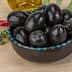 A bowl filled with black olives on a wooden surface with herbs and spices nearby. - Olive Oil Times