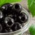 A close-up of black olives in a glass bowl with green leaves nearby. - Olive Oil Times
