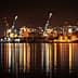 Night view of a port with illuminated shipping containers and cranes reflecting on the water. - Olive Oil Times