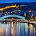 Illuminated bridge over a river with a cityscape and hillside in the background at night. - Olive Oil Times
