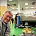 A man smiling in an olive oil processing facility with machinery and bottles of olive oil in the foreground. - Olive Oil Times