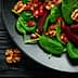A black plate filled with spinach leaves, pomegranate seeds, and walnuts arranged together. - Olive Oil Times
