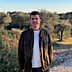 A man wearing a dark jacket and white sweater standing in an olive grove with trees in the background. - Olive Oil Times