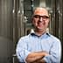 A man with glasses smiling and standing with arms crossed in a production facility surrounded by large metal tanks. - Olive Oil Times