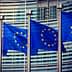 Four European Union flags displayed on poles in front of a modern building. - Olive Oil Times