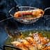 A metal strainer lifting a fried sausage from a pan of hot oil with bubbling surface. - Olive Oil Times