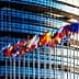 Multiple flags of European countries displayed in front of a modern glass building. - Olive Oil Times