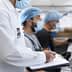 Three workers in a food processing facility wearing masks and hairnets, with one taking notes on a clipboard. - Olive Oil Times