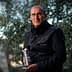 Man in a black jacket holding a bottle while standing among olive trees. - Olive Oil Times