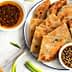 A plate of scallion pancakes arranged with two bowls of dipping sauces and green onions on a marble surface. - Olive Oil Times