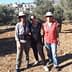 Three individuals standing together in an olive grove, wearing hats and casual clothing. - Olive Oil Times