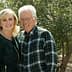 A couple smiling while posing outdoors in front of olive trees, wearing casual clothing. - Olive Oil Times