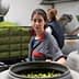 Woman sorting olives in a production facility with large containers in the background. - Olive Oil Times