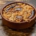 A baked dish containing sausages, onions, and grains in a round ceramic container on a wooden surface. - Olive Oil Times