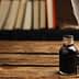 A glass ink bottle with a cork stopper and a quill pen resting in a holder on a wooden table. - Olive Oil Times