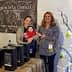 Two women and a baby at an olive oil stand with products displayed in a tent. - Olive Oil Times