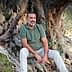 Man with a beard sitting on the roots of an olive tree, wearing a green shirt and light-colored pants. - Olive Oil Times